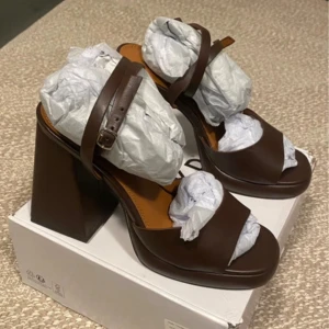 Bruna sandaler med klack - Snygga bruna sandaler med bred klack och justerbart spänne. Perfekta för en stilren look med öppen tå och elegant design.