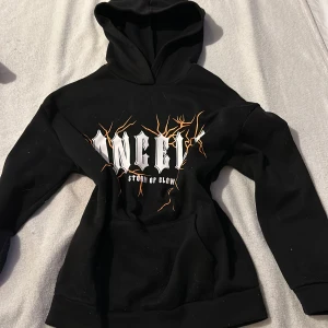 Svart hoodie med tryck från SHEIN - Cool svart hoodie från SHEIN med trycket 'ANGELS' och 'STORM OF GLOW' i vitt och orange. Hoodien har en klassisk design med huva och en stor ficka framtill. Perfekt för en avslappnad stil.