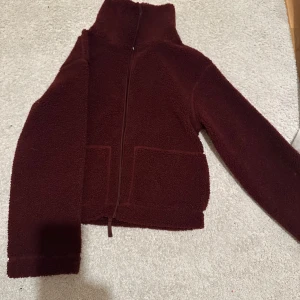 Vinröd teddyjacka - Mysig vinröd teddyjacka med dragkedja och hög krage. Perfekt för kyliga dagar med sin mjuka och varma känsla. Jackan har långa ärmar och en croppad passform som ger en trendig look. Prislappen kvar!