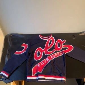 Ralph Lauren - Säljer en mörkblå tröja från Ralph Lauren med stora röda och vita detaljer. Tröjan har långa ärmar och en rund halsringning med randiga muddar i rött och vitt. Perfekt för en sportig look! Den är äkta och i bra skick.