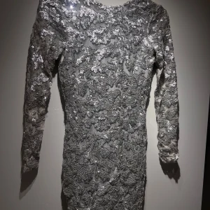 Glittrig silverklänning med paljetter - Rebecca Stella - Snygg och glittrig silverklänning täckt med paljetter. Den har långa ärmar och en öppen rygg som ger en elegant touch. 