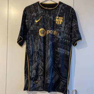 Svart och guld Barcelona fotbollströja från Nike - Snygg svart Barcelona fotbollströja med guldiga detaljer och klubbens emblem. Tröjan har korta ärmar och ett unikt mönster som täcker hela plagget. Perfekt för fans som vill visa sitt stöd med stil. Special edition 23/24