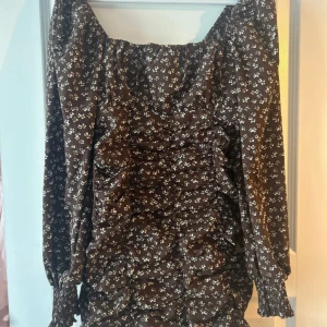 Brun blommig klänning - Charmig brun klänning med små vita blommor och volangdetaljer. Klänningen har långa ärmar och en smickrande rynkad design framtill. Perfekt för en stilfull look.