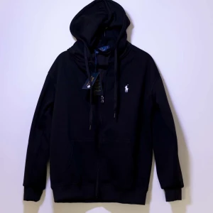 Ralph Lauren tröja - Helt ny Ralph Lauren tröja med dragkedja och det klassiska logotypbroderiet på bröstet. Perfekt för en stilren och bekväm look. Den har en justerbar huva med snören.