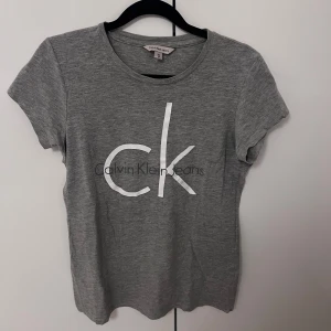 Grå t-shirt från Calvin Klein - Snygg grå t-shirt från Calvin Klein med det ikoniska 'ck' trycket på framsidan. Perfekt för en avslappnad stil. Tillverkad i mjukt material för extra komfort. 120kr
