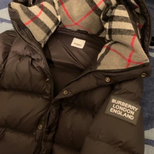 Svart pufferjacka från Burberry - Snygg svart pufferjacka från Burberry med klassiskt rutigt foder i huvan. Jackan har dragkedja och knappar framtill samt två praktiska fickor med dragkedja. Perfekt för kyliga dagar. Man kan också göra jackan till en väst ifall man skulle vilja det. Pris kan diskuteras!