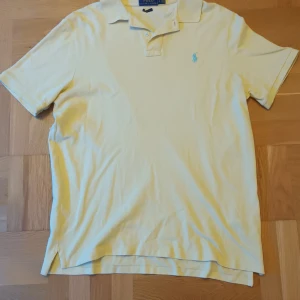 Gul pikétröja från Ralph Lauren - Säljer en klassisk gul pikétröja från Ralph Lauren med korta ärmar och en broderad logga på bröstet. Perfekt för en avslappnad och stilren look. Tröjan har en krage med knappar.