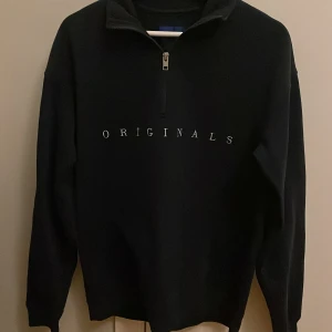 Svart tröja från Jack & Jones - Snygg svart tröja från Jack & Jones med texten 'ORIGINALS' på bröstet. Tröjan har en dragkedja vid halsen och långa ärmar. Perfekt för en stilren och avslappnad look.