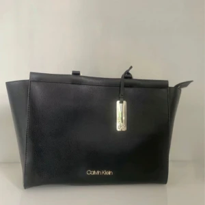 Svart handväska från Calvin Klein - Elegant svart handväska från Calvin Klein i syntetmaterial. Väskan har en stilren design med dragkedja och ett dekorativt metallspänne framtill. Perfekt för att bära dina dagliga nödvändigheter med stil.