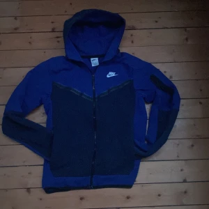 Blå och svart nike tech fleec jättebra skick  - Snygg blå och svart hoodie från Nike med dragkedja och huva. Perfekt för en sportig look eller avslappnade dagar. Den har en praktisk ficka med dragkedja på ärmen.