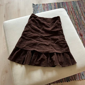 Brun manchesterkjol med volang - Snygg brun manchesterkjol med volangdetalj. Kjolen har en mellankort längd och är perfekt för höst och vinter. Den har en bekväm passform och är tillverkad i ett mjukt material.