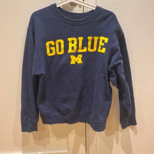 Mörkblå sweatshirt från Fruit of the Loom - Säljer en mörkblå Michigan State sweatshirt från Fruit of the Loom med texten 'GO BLUE' i gult på framsidan. Tröjan har långa ärmar och en rund halsringning. Perfekt för en avslappnad stil. Tröjan är relativt cropped och är ganska kort i passformen.