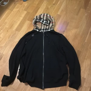 Svart huvtröja från Burberry - Säljer en stilren svart huvtröja från Burberry med dragkedja och klassiskt rutig huva. Tröjan är tillverkad i bomull och har långa ärmar. Perfekt för en avslappnad och trendig look. Köpt för drygt en månad sedan ny pris ca 9000kr