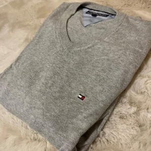 Grå tröja från Tommy Hilfiger - Säljer en stilren grå tröja från Tommy Hilfiger med v-ringning och det klassiska logotypmärket på bröstet. Tröjan är tillverkad i bomull och har långa ärmar med ribbade muddar. Perfekt för en avslappnad och snygg look.