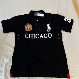 Svart pikétröja från Ralph Lauren - Snygg svart pikétröja från Ralph Lauren med 'Chicago' tryck och broderad logga. Tröjan har korta ärmar och nummerdetaljer på ärmarna och ryggen. Perfekt för en sportig look!