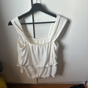 Vit ribbad topp - Säljer en stilren vit ribbad topp med breda axelband och volangdetaljer. Perfekt för en somrig look. Toppen har en luftig passform och är tillverkad i ett mjukt material. Den är från Shein!