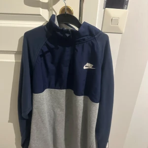Blå och grå hoodie från Nike - Säljer en snygg blå och grå hoodie från Nike med stor logga på bröstet och ärmen. Perfekt för en sportig look eller avslappnad stil. Hoodien har en dragkedja och är tillverkad i mjukt material för extra komfort.