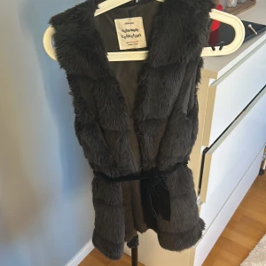 Svart pälsväst från Zara - Snygg svart pälsväst från Zara Girls, perfekt för att hålla sig varm och stilren. Västen har ett bälte i midjan för en figurnära passform och är en del av deras Outerwear Collection.