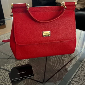 Röd handväska från Dolce & Gabbana - Elegant röd handväska från Dolce & Gabbana i skinn med guldfärgade detaljer. Väskan har en avtagbar axelrem och en stilren design med en logotypplatta på framsidan. Perfekt för att ge en färgklick till din outfit.