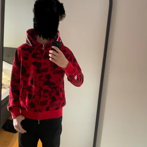 Röd camo hoodie med dragkedja - Säljer en cool röd camo hoodie med dragkedja och hajmotiv på huvan. Den har långa ärmar och en unik design med broderade detaljer. Perfekt för dig som vill sticka ut! M passar S.