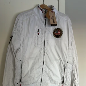 Vit jacka från Deeluxe - Vit jacka från Deeluxe med broderad detalj på bröstet. Jackan har en hög krage med randig kant och flera praktiska fickor.  Storlek på jackan är XL men känns som L