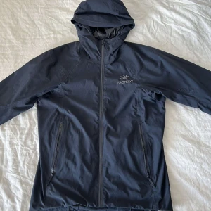 Arcteryx - Säljer en stilren svart vindjacka från Arc'teryx med huva och dragkedja. Perfekt för blåsiga dagar och utomhusaktiviteter. Jackan har två praktiska fickor med dragkedja framtill.