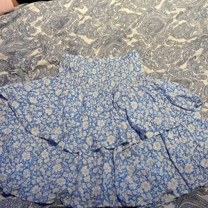 Blommig volangkjol - Charmig kort blommig kjol i blått och vitt med volanger. Perfekt för en somrig look med elastisk midja för bekväm passform.