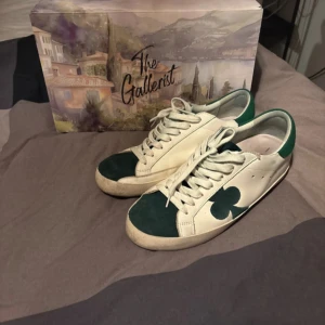 Vita och gröna sneakers från The Gallerist - Snygga vita sneakers med gröna detaljer från The Gallerist. Skorna har en grön klöverdetalj på sidan och grönt parti vid hälen. Perfekta för en stilren look. Skicka för fler bilder. Dem är i sjukt bra skick. Priset är ej hugget i sten. Box medföljer.