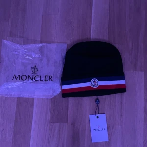 Svart mössa från Moncler - Snygg svart mössa från Moncler med röd, vit och blå randdetalj. Mössan har Monclers logotyp framtill och är perfekt för att hålla dig varm med stil. Mycket snygg mössa. Perfket för kyliga dagar! Helt ny aldrig använda! Pris kan diskuteras vid snabb affär!