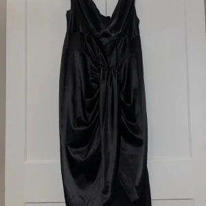 Svart långklänning i satin - Elegant svart långklänning i satin med v-ringad design och draperad detalj framtill. Perfekt för en festlig kväll.