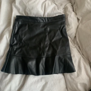 Svart kjol från Kappahl - Snygg svart kjol i skinnimitation från Kappahl. Kjolen har en stilren design med en lätt volangdetalj vid fållen. Perfekt för att ge en edgy touch till din outfit.