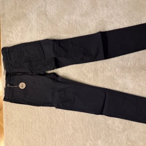 Svarta kostymbyxor från East west - Eleganta svarta kostymbyxor med dragkedja och knappstängning. Perfekta för en stilren look. Byxorna har en slim passform och är tillverkade i ett bekvämt material. Storlek 30/32. Helt ny med prislapp kvar. Orginalpris 800kr.