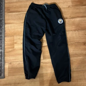 Mörkblåa träningsbyxor från Manchester City - Snygga mörkblåa träningsbyxor med Manchester City-logga. Byxorna har en elastisk midja med snörning och ljusblåa detaljer längs sidorna. Perfekta för fotbollsträning eller avslappnade dagar. Pris kan diskuteras 