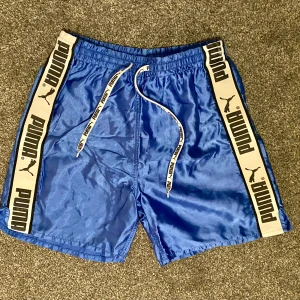 Blå shorts från Puma - Retro blå shorts från Puma med vita detaljer. Shortsen har en midja med dragsko. De är gjorda i ett glansigt och bekvämt material. Märkta storlek 164 eller XS, se bilder för mått.