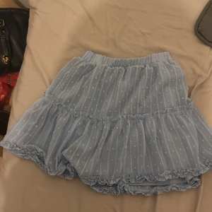 Ljusblå kortkjol med volang - Söt ljusblå kortkjol med volangdetaljer och ett diskret prickigt mönster. Kjolen har en elastisk midja för bekväm passform och är perfekt för varma dagar.