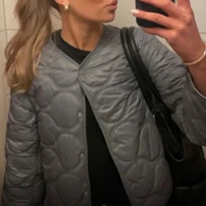 Grå quiltad jacka från mangon  - Helt sprillans ny (lappen sitter kvar) snygg grå quiltad jacka från mango med rund hals och tryckknappar. Perfekt för lager-på-lager under kyliga dagar. Jackan har en stilren design med långa ärmar och en lätt glansig finish. (Köpt för 570kr)