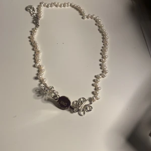 Pärlhalsband med silverdetaljer från snö - Elegant pärlhalsband med vita pärlor och dekorativa silverdetaljer. Halsbandet har en unik design med en rund, mörk detalj i mitten. Perfekt för att ge en touch av klass till din outfit.