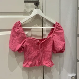 Rosa croppad topp med puffärmar - Söt rosa croppad topp med puffiga kortärmar och smockdetaljer. Toppen har en knytning framtill och volangkant nedtill, perfekt för en somrig look.
