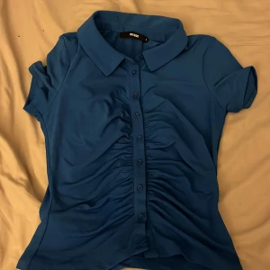 Blå kortärmad skjorta från Bik Bok - Snygg blå skjorta från Bik Bok med korta ärmar och knappar framtill. Skjortan har en klassisk krage och rynkade detaljer längs knappraden som ger en unik touch. Perfekt för en stilren look.