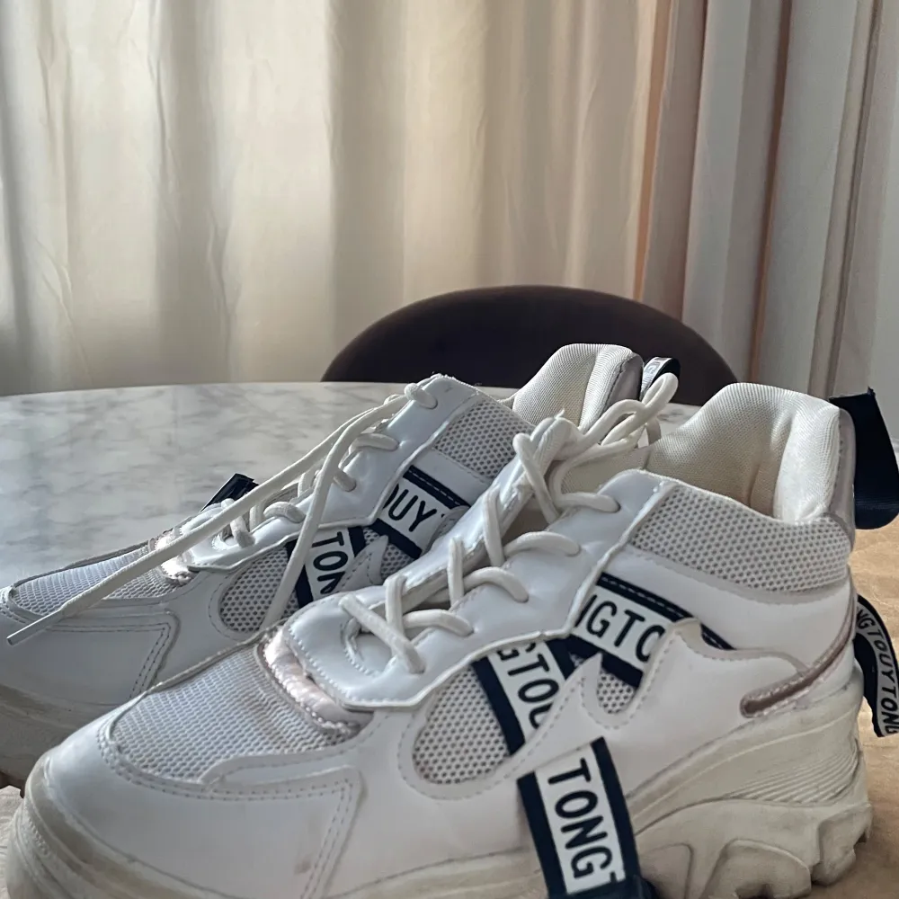 Snygga vita sneakers med tjock sula och textdetaljer i svart och vitt. Skorna har snörning och en modern design som passar perfekt för en trendig look.. Kengät.