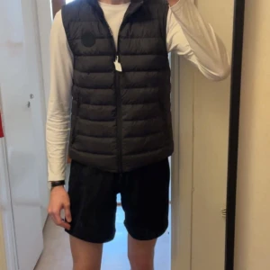 Svart dunväst från Woolrich - Säljer en stilren svart dunväst från Woolrich. Västen har en dragkedja framtill och två praktiska fickor med dragkedjor. Perfekt för lager-på-lager under kyliga dagar.