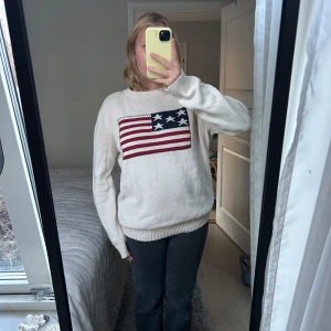 Vit stickad tröja med amerikansk flagga - Säljer en vit stickad tröja med en stor amerikansk flagga på bröstet. Tröjan har långa ärmar och en bekväm passform. Perfekt för en avslappnad stil.