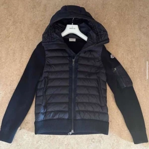 Svart jacka från Moncler - Snygg svart jacka från Moncler med quiltad design och huva. Jackan har dragkedja framtill och långa ärmar. Perfekt för kyligare dagar. Kvitto finns storlek 1/S