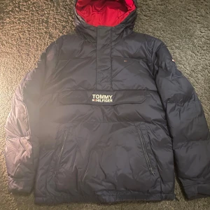 Navy Blue dunjacka från Tommy Hilfiger - Säljer en snygg Navy Blue dunjacka från Tommy Hilfiger med röd insida. Jackan har en stor ficka framtill med dragkedja och en huva för extra värme. Perfekt för kalla vinterdagar! 