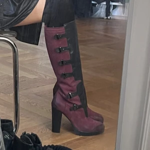 Vinröda och svarta stövlar med klack - Storlek: 39. Snygga vinröda stövlar med svarta detaljer och hög klack. De har en cool design med flera spännen och dragkedja på sidan. Perfekta för höst och vinter, och ger en edgy touch till vilken outfit som helst. Passar både till vardags och fest!