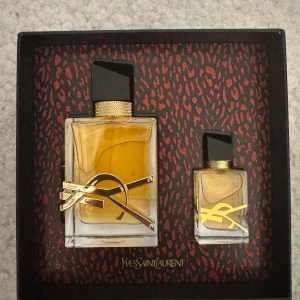 Yves Saint Laurent Libre parfymset - Oanvänd YSL Elegant parfymset från Yves Saint Laurent med två flaskor av doften Libre. Flaskorna har en stilren design med svart lock och guldiga detaljer. Förpackningen har ett djärvt mönster i rött och svart med YSL-loggan i guld.