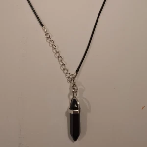 Svart halsband med kristallhänge - Snyggt svart halsband med ett coolt kristallhänge i silverinfattning. Perfekt för att ge din outfit en mystisk touch. Kedjan är justerbar och passar de flesta storlekar. Kommer i en praktisk förpackning, redo att användas eller ges bort som present.
