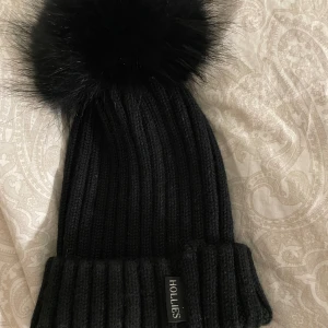 Svart ribbad mössa med pälsboll från Hollies - Snygg svart ribbad mössa från Hollies med en stor fluffig pälsboll på toppen. Perfekt för att hålla sig varm och stilren under kalla dagar. Mössan har en uppvikt kant och är i ett mjukt och bekvämt material. Säljer för den desvärre inte kommer till användning:/ köparen står för frakt:)  