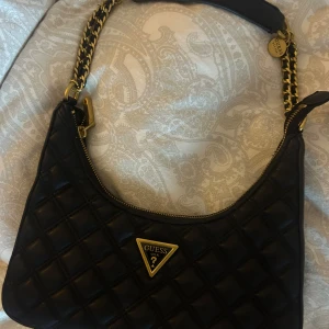 Svart axelväska från Guess - Säljer en snygg svart axelväska från Guess, självklart äkta. Väskan har en kedjedetalj på axelremmen och en dragkedja för säker förvaring. Perfekt för både vardag och fest! 💗