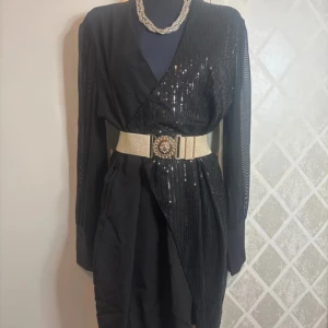 Svart glittrig klänning med bälte - Elegant svart klänning med glittriga paljettdetaljer. Bältet ingår ej!  Klänningen har långa ärmar i ett transparent material och en omlott-design framtill.